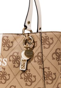 Tan handbag with a patterned exterior, featuring a gold chain and keychain accent, and white leather handles. Brand logo visible.