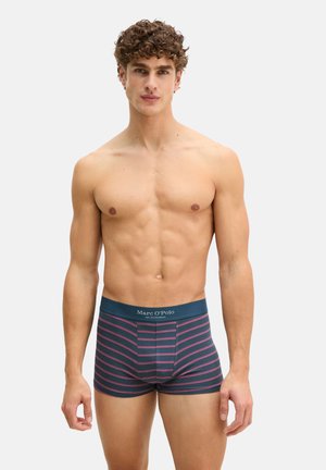 Athletischer Mann mit lockigem Haar, der marineblaue Boxer-Shorts mit rosa Streifen und dem Marc O'Polo-Logo am Bund trägt, vor einem einfarbigen Hintergrund stehend.