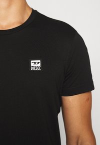 Nahaufnahme einer Person, die ein schwarzes Diesel-T-Shirt trägt, mit einem kleinen weiß-schwarzen Diesel-Logo-Patch auf der linken Brust.