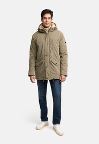 Ein Mann steht mit einer beigen Kapuzen-Winterjacke, blauen Jeans und weißen Turnschuhen vor einem einfarbigen Hintergrund.