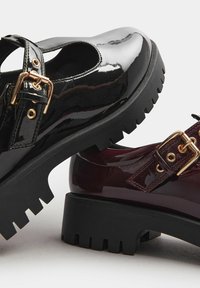 Un par de zapatos de plataforma: uno en cuero negro brillante con una correa y una hebilla dorada, el otro en burdeos brillante con elementos de diseño similares.