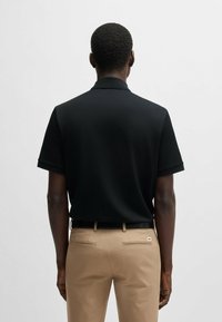 Black polo shirt with short sleeves, textured fabric, and a collar, paired with beige trousers featuring back pockets and a logo tag.