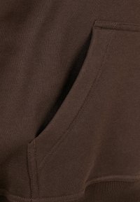 Tejido de sudadera marrón con bolsillo tipo canguro, textura suave y dobladillo acanalado. Detalle de costura visible a lo largo del bolsillo y los bordes.