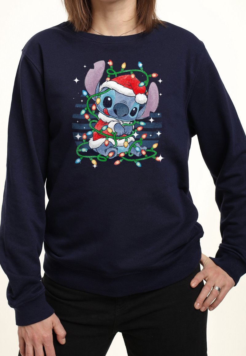 Personnage Stitch portant un bonnet de Père Noël et enveloppé de guirlandes lumineuses colorées sur un t-shirt à manches longues bleu marine porté par une personne.