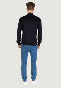 Hombre de espaldas vistiendo un suéter negro de manga larga, jeans azules y zapatillas blancas frente a un fondo liso.