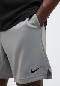 Άτομο με σκουρόχρωμο δέρμα φοράει ανοιχτογκρι σορτς Nike με μαύρο λογότυπο, το χέρι στην δεξιά τσέπη και μαύρο πουκάμισο μερικώς ορατό.