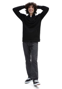 Felpa nera con zip e tasca a marsupio, abbinata a pantaloni grigi e sneakers nere. Il modello sta in piedi con le braccia sollevate in una posa sorridente.