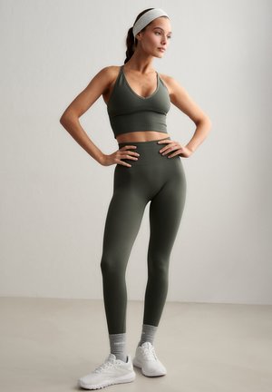 Kvinde i olivengrøn sports-bh og leggings står med hænderne på hofterne, iført grå sokker, hvide sneakers og et hvidt pandebånd.