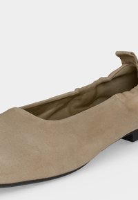 Απαλό μπεζ σουέτ επίπεδο παπούτσι με ραβδωτή ελαστική γραμμή και χαμηλό μαύρο τακούνι, εμφανιζόμενο από πλάγια γωνία σε λευκό φόντο.