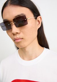 Rectangle sunglasses with dark lenses, gold accents, and clear detail near hinges, worn with a white t-shirt featuring a red graphic.