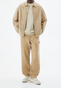 Mężczyzna w beżowej kurtce, jasnobeżowym swetrze, luźnych beżowych spodniach i białych sneakerach, stojący z rękami w kieszeniach na białym tle.