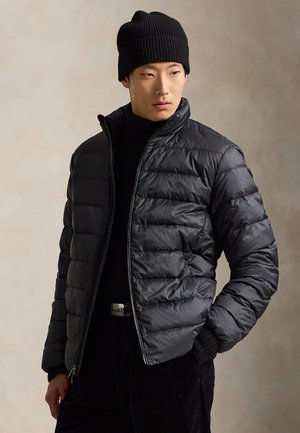 Homme portant un bonnet en maille noir, une doudoune noire zippée, un col roulé noir et un pantalon noir, debout devant un fond beige.