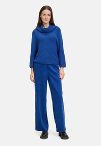 Niebieski sweter dzianinowy z dekoltem w czworobok, zestawiony z niebieskimi spodniami z sztruksu o szerokich nogawkach. Całość dopełniają czarne botki za kostkę.