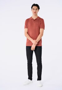 Polo shirt rosso a maniche corte con finitura testurizzata, abbinato a jeans neri slim-fit e sneakers bianche, in piedi contro uno sfondo chiaro.