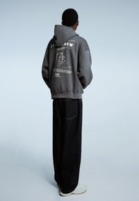 Grijze hoodie met capuchon en "COLD BREW" opdruk op de achterkant, gecombineerd met losse zwarte jeans en lichte sneakers. Minimalistisch ontwerp.