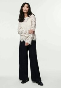 Lace long-sleeve top in off-white with a scalloped hem, paired with wide-leg black pants and black shoes. Textured fabric with intricate patterns.