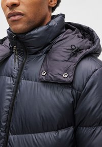 Casaco acolchoado azul-marinho com gola alta e capuz, apresentando um fecho de correr frontal e ilhós metálicos, textura lisa com acolchoamento matelassado.