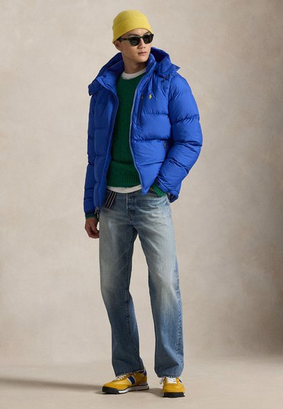 Modra puhasta jakna s kapuco, zelena pletena majica, svetlo modre kavbojke in rumena kapa. Obutev ima rumene in modre poudarke.