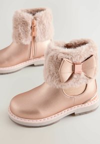 Botas tobilleras color oro rosa con un puño de piel sintética y detalle de lazo. Cuentan con un zipper lateral, suela ligera y textura suave.