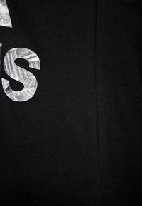 Černá látka mikiny s výraznými, texturovanými bílými písmeny. Materiál působí měkce s viditelným švem na boku.