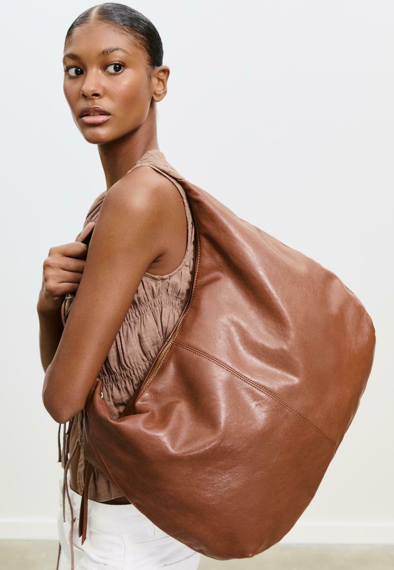 Brown leather shoulder bag with a soft, rounded shape and visible stitching. Model holds the bag with a loose, textured top.