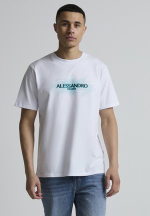 Giovane uomo con capelli corti che indossa una maglietta bianca Alessandro Milano e jeans blu, in piedi davanti a uno sfondo grigio uniforme.