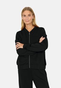 Woman with long blonde hair wears black zip-up jacket and matching black pants, standing with arms crossed against plain background.