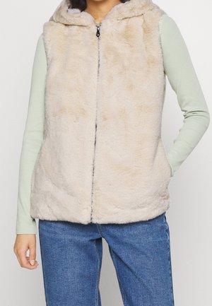 Femme portant une veste sans manches à capuche beige clair et duveteuse par-dessus un haut à manches longues vert pâle et un jean bleu.