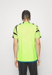 Camiseta deportiva de color verde neón con mangas cortas, detalles en negro y un patrón de puntos en los hombros, con un cuello redondo y ribetes laterales azules.