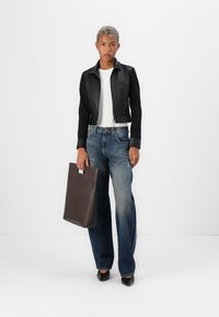 Giacca cropped in pelle nera, camicia bianca, jeans svasati blu sbiaditi, tiene una borsa tote marrone scuro e indossa scarpe nere eleganti.