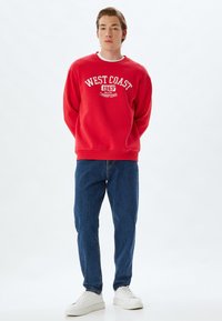 Sudadera roja con el texto blanco "CAMPEONES UNIVERSITARIOS DE LA COSTA OESTE 1965", combinada con jeans azules y zapatillas blancas. Diseño clásico de cuello redondo.