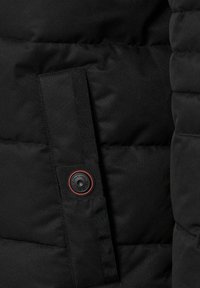 Dettaglio di una giacca nera trapuntata che mostra una tasca a patta verticale con un bottone a pressione nero con rifiniture rosse e il testo "CASACMODA".