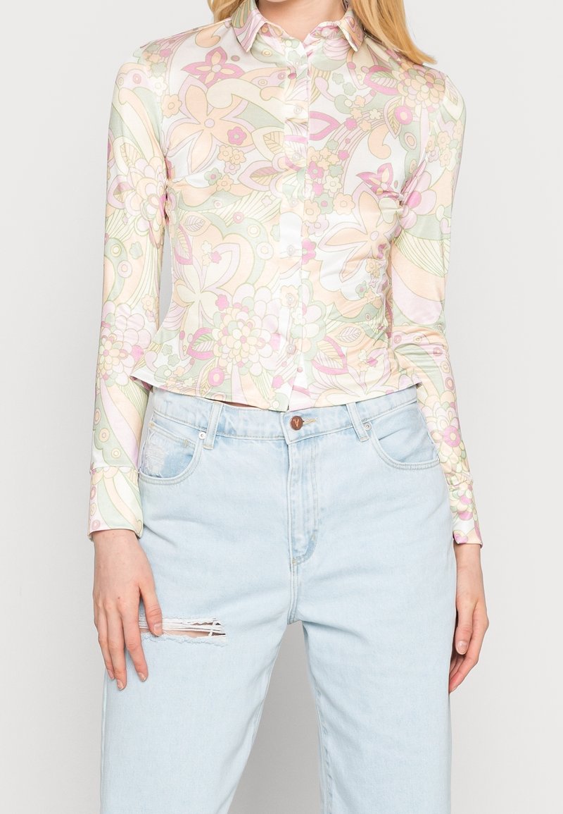 Mujer vestida con una blusa abotonada de manga larga con estampado floral en tonos pastel, combinada con unos pantalones vaqueros de tiro alto de color azul claro, con un pequeño desgarro en el muslo.