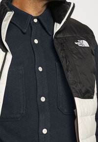 Vadderad väst i svart och vitt, med dragkedja fram, ton-i-ton sömnad och logotyp; bärs över en mörkblå strukturerad skjorta med knappar.