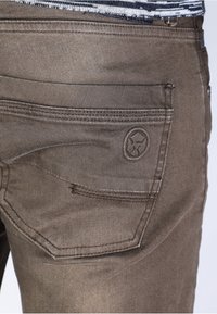 Jeans de mezclilla marrones con un bolsillo trasero con detalle del logo, acentos cosidos y un acabado de tela texturizada.