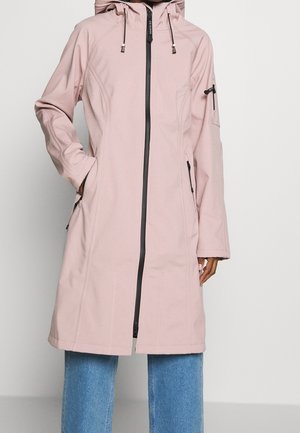 Long manteau à capuche rose clair avec fermeture éclair noire à l'avant, poches latérales zippées et poche sur la manche, porté sur un jean bleu.