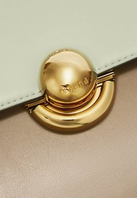Kremowa i beżowa skórzana torebka z okrągłym, złotym zapięciem z wytłoczonym logo. Gładka tekstura z kontrastującymi kolorami.