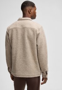 Giacca in pile color beige, testurizzata, con colletto, polsini con bottoni e dettagli delle cuciture. Il design è a vestibilità rilassata e arriva fino ai fianchi.