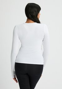 Langärmliges, weißes Sportoberteil mit strukturiertem Stoff, taillierter Passform und Daumenlöchern an den Ärmelenden, kombiniert mit gemusterten schwarzen Leggings.