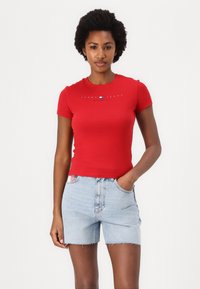 Rød kortermet t-skjorte i mykt materiale med «TOMMY JEANS» trykk i hvitt. Kombinert med lyseblå denimshorts med slitte kanter.