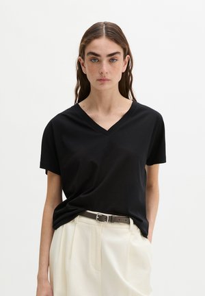 Frau mit langen braunen Haaren, die ein schwarzes T-Shirt mit V-Ausschnitt trägt, in hoch taillierte cremefarbene Hosen mit braunem Gürtel gesteckt.
