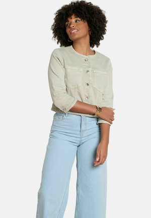 Woman with curly hair wearing a light beige buttoned jacket and high-waisted light blue wide-leg jeans, posing with one arm crossed.
