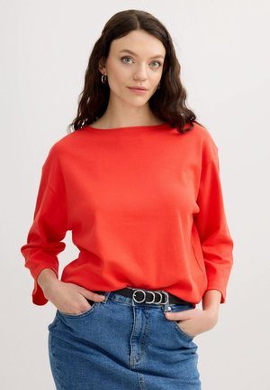 Mujer con cabello castaño ondulado, que lleva pendientes de aro dorados, una camiseta de manga larga roja brillante y vaqueros azules con un cinturón negro, manos en los bolsillos.