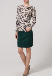 Top beige a maniche lunghe con stampa floreale nera, abbinato a una gonna di jeans verde scuro. La modella indossa scarpe con tacco beige.