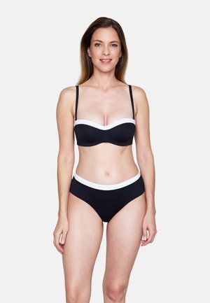 Vrouw staande, naar voren gericht, gekleed in een zwarte bikini met witte rand, glimlachend tegen een effen lichte achtergrond.
