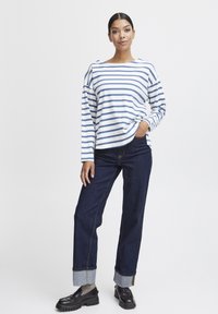 Long-sleeve striped shirt in white and blue, paired with dark denim jeans featuring a rolled cuff. Model wears black chunky shoes.