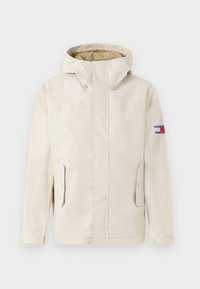 Chaqueta con capucha beige, con bolsillos frontales y cierres de solapa, mangas largas y un pequeño parche de logo rectangular en la parte superior de la manga izquierda.
