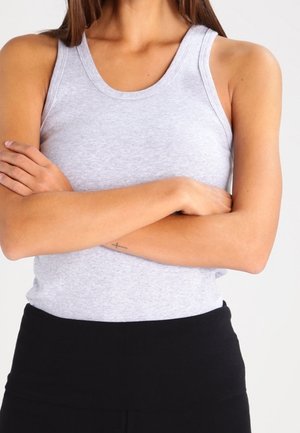 Personne portant un haut gris sans manches, les bras croisés, montrant un petit tatouage de croix à l'intérieur du bras.