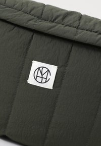 Donkergroene gevoerde tas met een geborduurd patroon, voorzien van een wit etiket met een zwart logo. Het materiaal lijkt lichtgewicht en duurzaam te zijn.