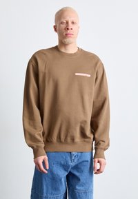 Sudadera de algodón marrón con cuello redondo, que presenta un pequeño logo rosa en el pecho, combinada con pantalones cortos de mezclilla azul.